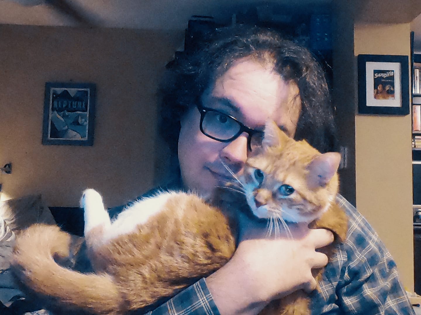 A man with 8cm long dark hair sits on a computer chair, holding an orange cat like a baby"